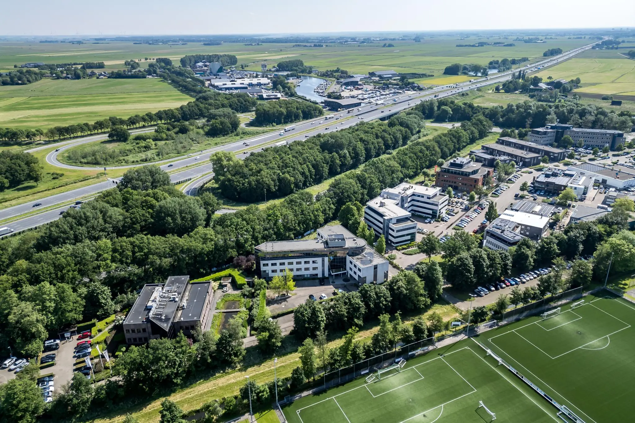 Luchtfoto van de Baarnsche Dijk met kantoorgebouwen, sportvelden en snelwegen omgeven door groen en weilanden.